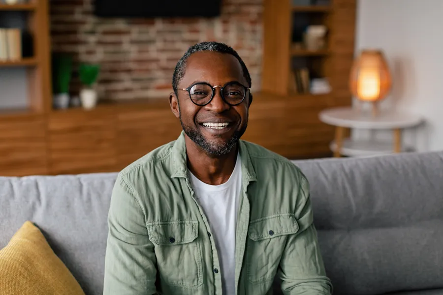 Andropause-Doctor A middle aged black man in a green button up shirt sits on his couch smiling, happy with his treatment for andropause and low testosterone from Julie Kane, NP of Trua Health & Wellness in Alaska.