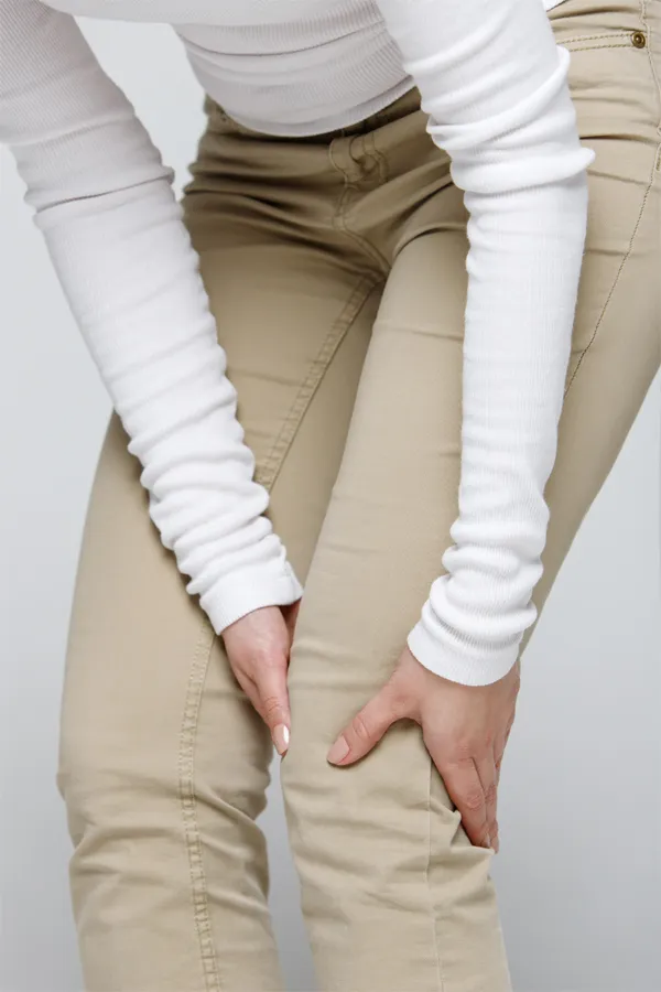 Arthritis-Treatment A woman wearing a white long-sleeve shirt and tan pants, clutching her knee, suffering from arthritis. Get arthritis treatment from Julie Kane, NP of Trua Health & Wellness in Alaska.