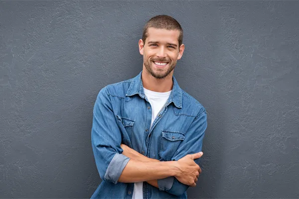 Hormones-for-Men-Doctor A man in a denim shirt stands smiling against a gray-blue wall, pleased with his testosterone hormone therapy from Julie Kane, NP of Trua Health & Wellness in Alaska.