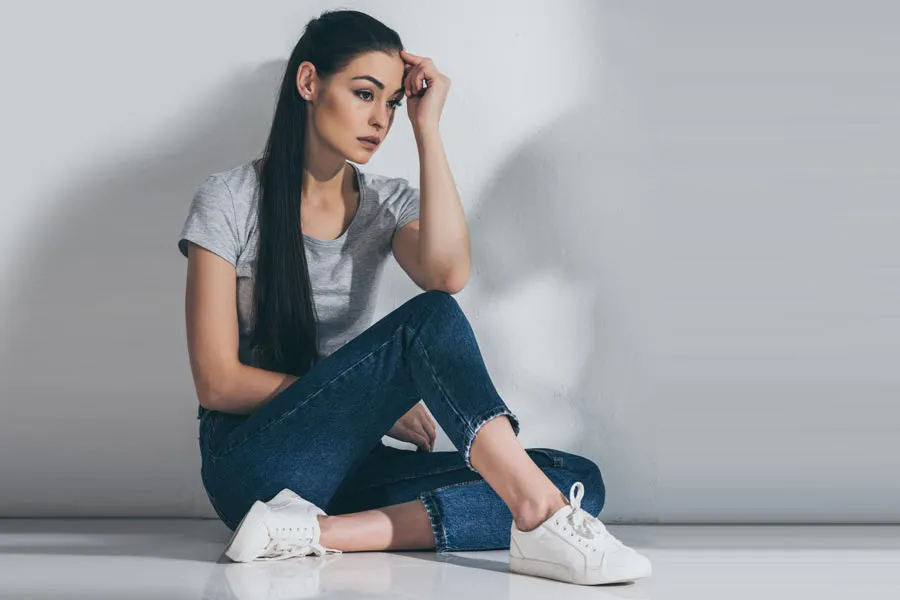 PCOS-Doctor A young brunette woman in a gray t-shirt and jeans sits against a gray wall with her head resting on her hand and a distressed expression. Get help for Polycystic Ovarian Syndrome (PCOS) from Julie Kane, NP of Trua Health & Wellness in Alaska.