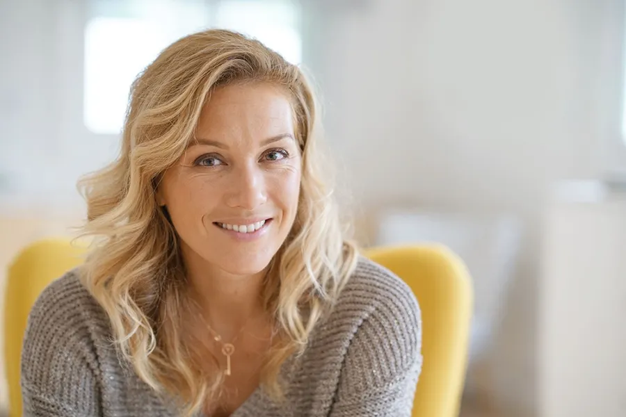 Perimenopause-Doctor A blonde woman wearing a a gray sweater sits inside in a yellow chair, smiling and happy with her perimenopause treatment from Julie Kane, NP of Trua Health & Wellness in Alaska.