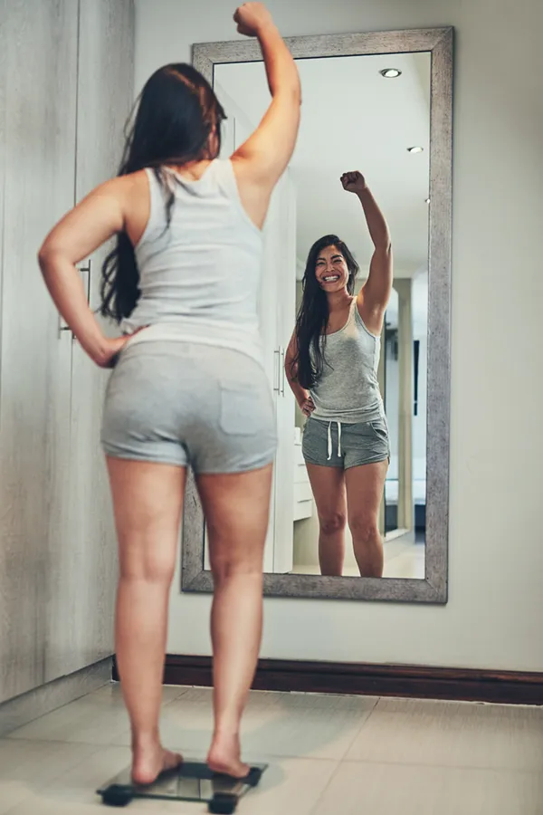 Tirzepatide-Treatment A woman in workout clothes stands on a scale looking in the mirror and pumping her hands in celebration of her successful Tirzepatide treatment from Julie Kane, NP of Trua Health & Wellness in Alaska.