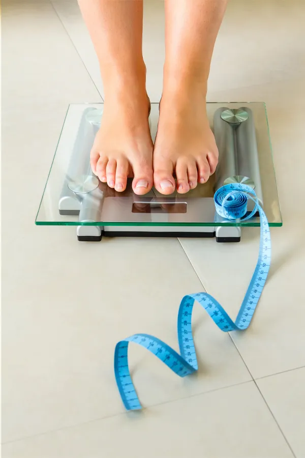 Weight-Loss-Resistance-Treatment Close-up of a woman's feet standing on a scale, with measuring tape by her toes, getting treatment for weight loss resistance from Julie Kane, NP of Trua Health & Wellness in Alaska.